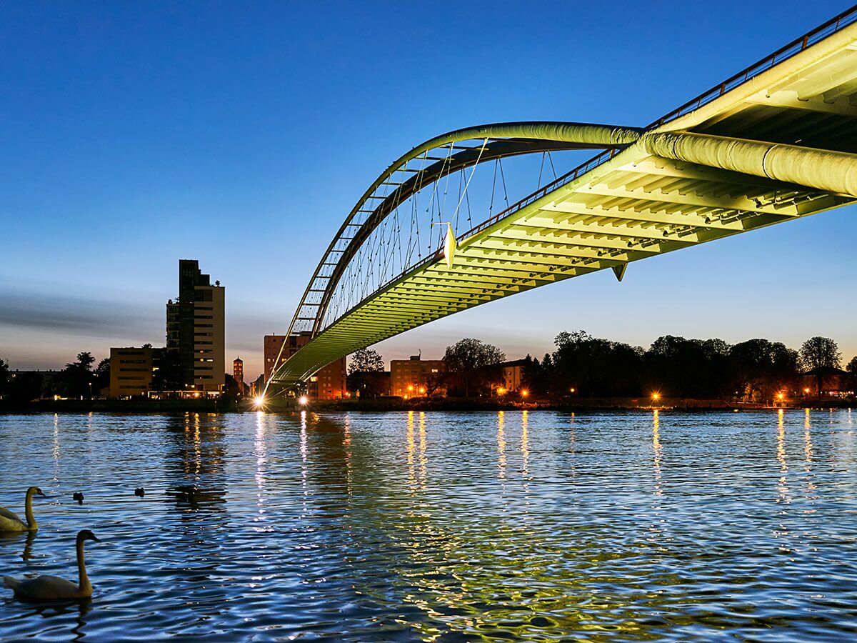 Beleuchtete Bogenbrücke über einen Fluss bei Nacht mit Stadtkulisse im Hintergrund und Schwänen im Wasser.