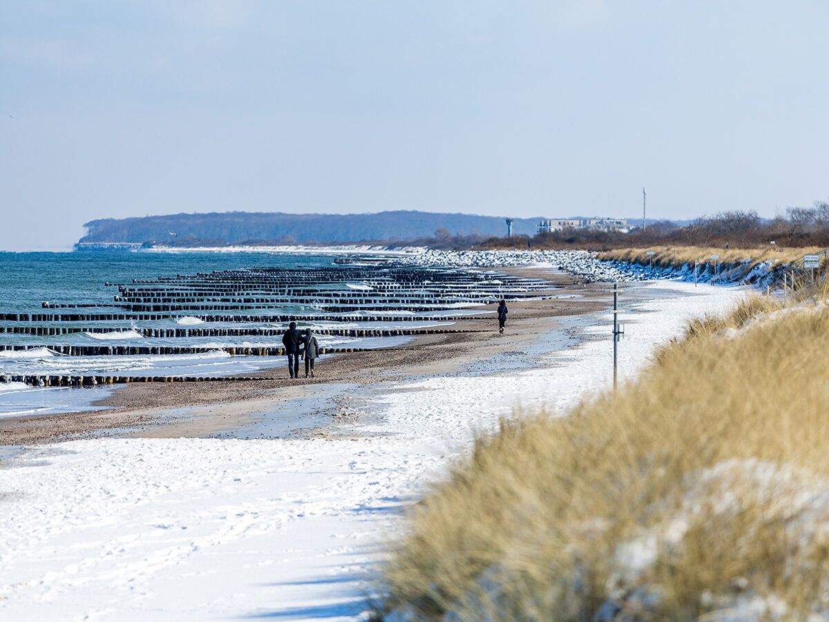 Schneebedeckter Ostseestrand mit Holz-Buhnen, wo Menschen im Winter spazieren gehen. Im Hintergrund eine schneebedeckte Klippe.