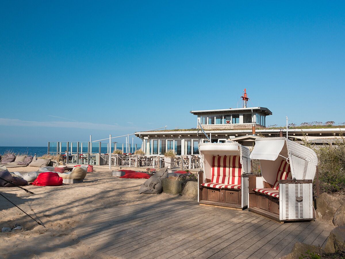 Zwei gestreifte Strandkörbe auf Holzdeck vor Sandstrand, bunten Sitzsäcken und Restaurant am Meer unter blauem Himmel.