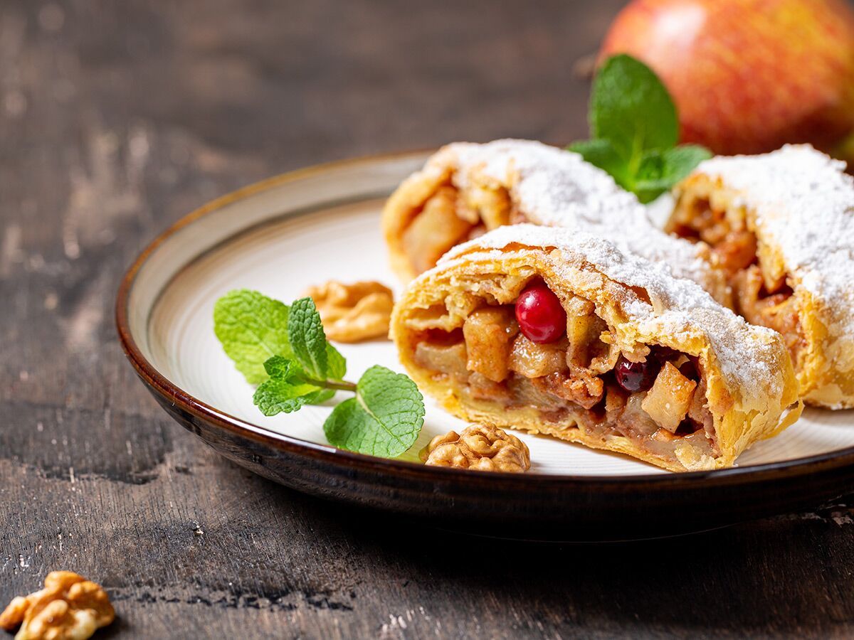 Scheiben Apfelstrudel mit Puderzucker, Minze, Preiselbeeren und Walnüssen auf Holztisch, im Hintergrund Apfel.