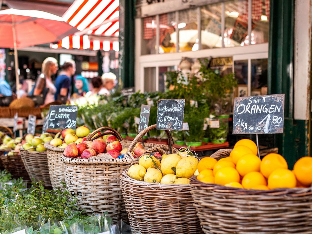 Bunter Obstmarkt mit Körben voller Äpfel, Zitronen und Orangen, Preisschilder und unscharfe Personen im Hintergrund.