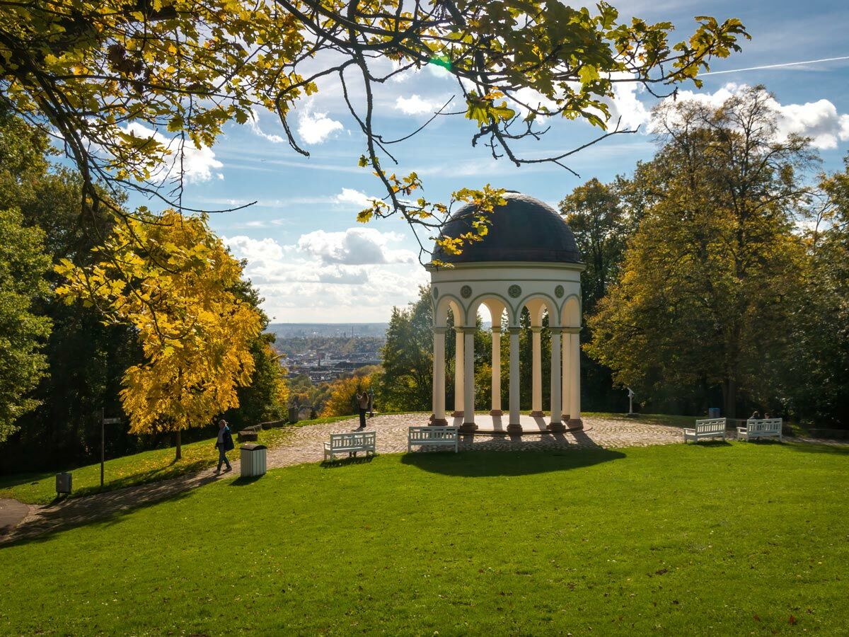 Herbstlicher Park mit klassischem Pavillon, gelben Bäumen, Menschen und Weitblick auf eine Stadt unter blauem Himmel.