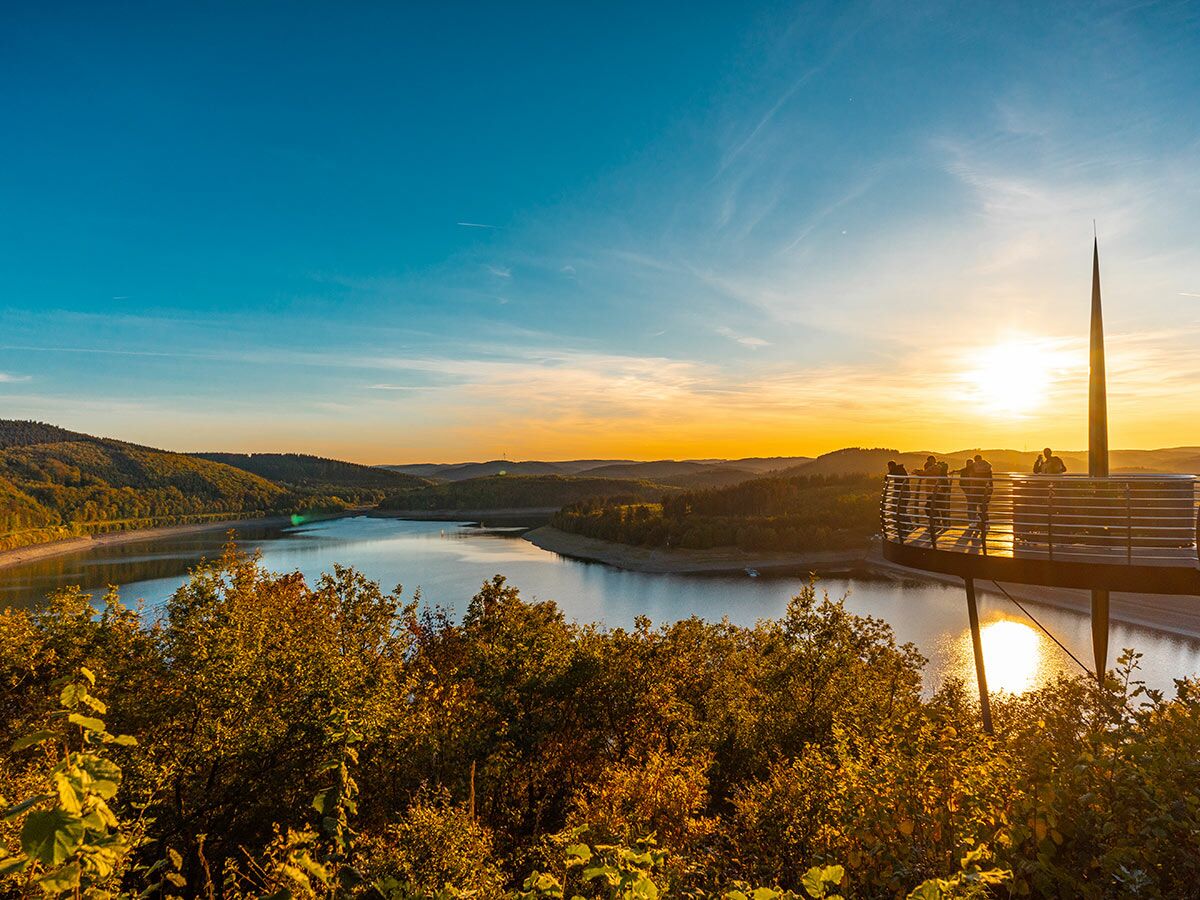 Eine erhöhte Aussichtsplattform mit Menschen blickt auf einen großen See und bewaldete Hügel bei Sonnenuntergang.