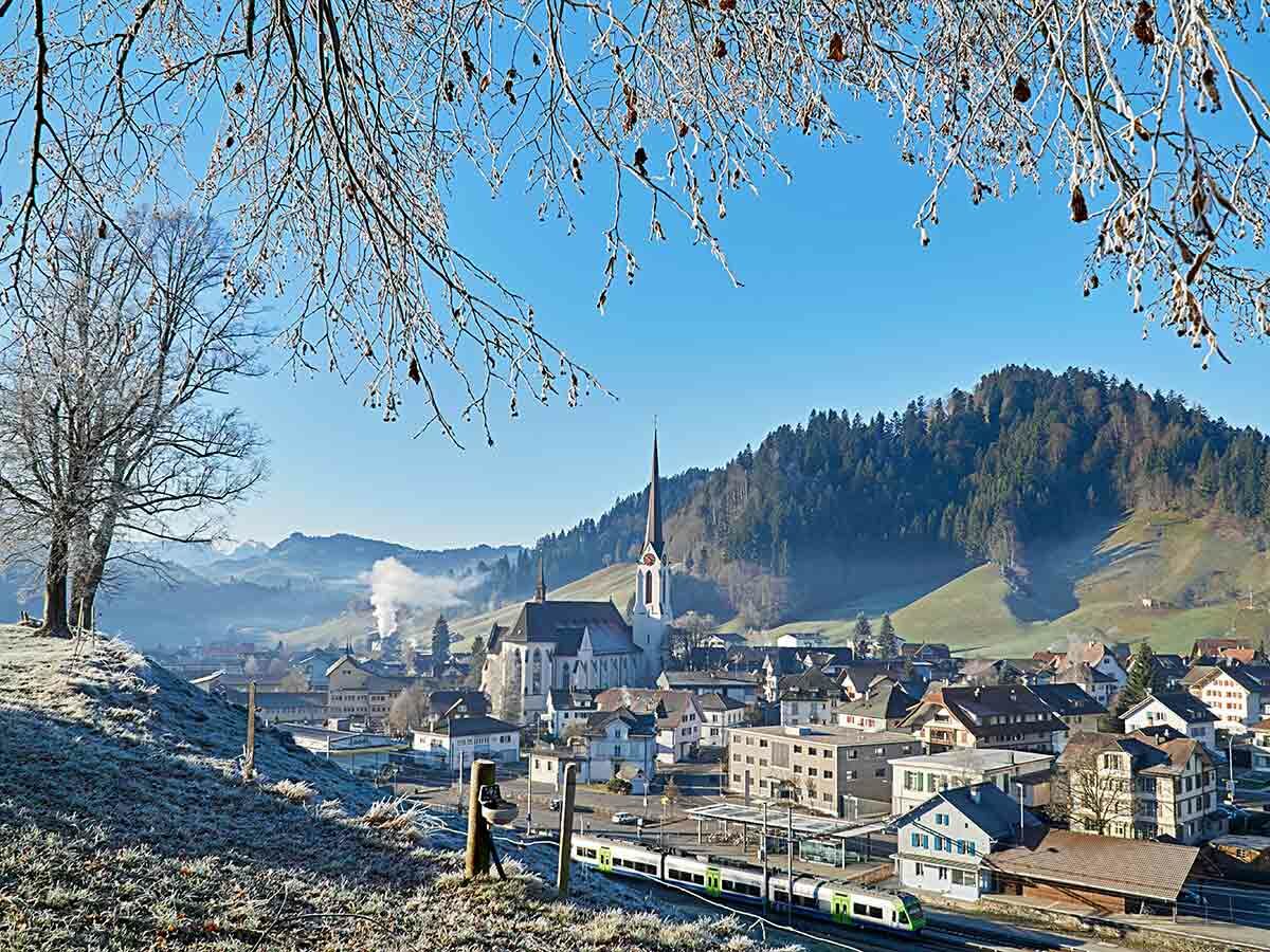 Winterliches Dorf in den Schweizer Alpen mit Kirche, frostiger Landschaft und einem vorbeifahrenden Zug.