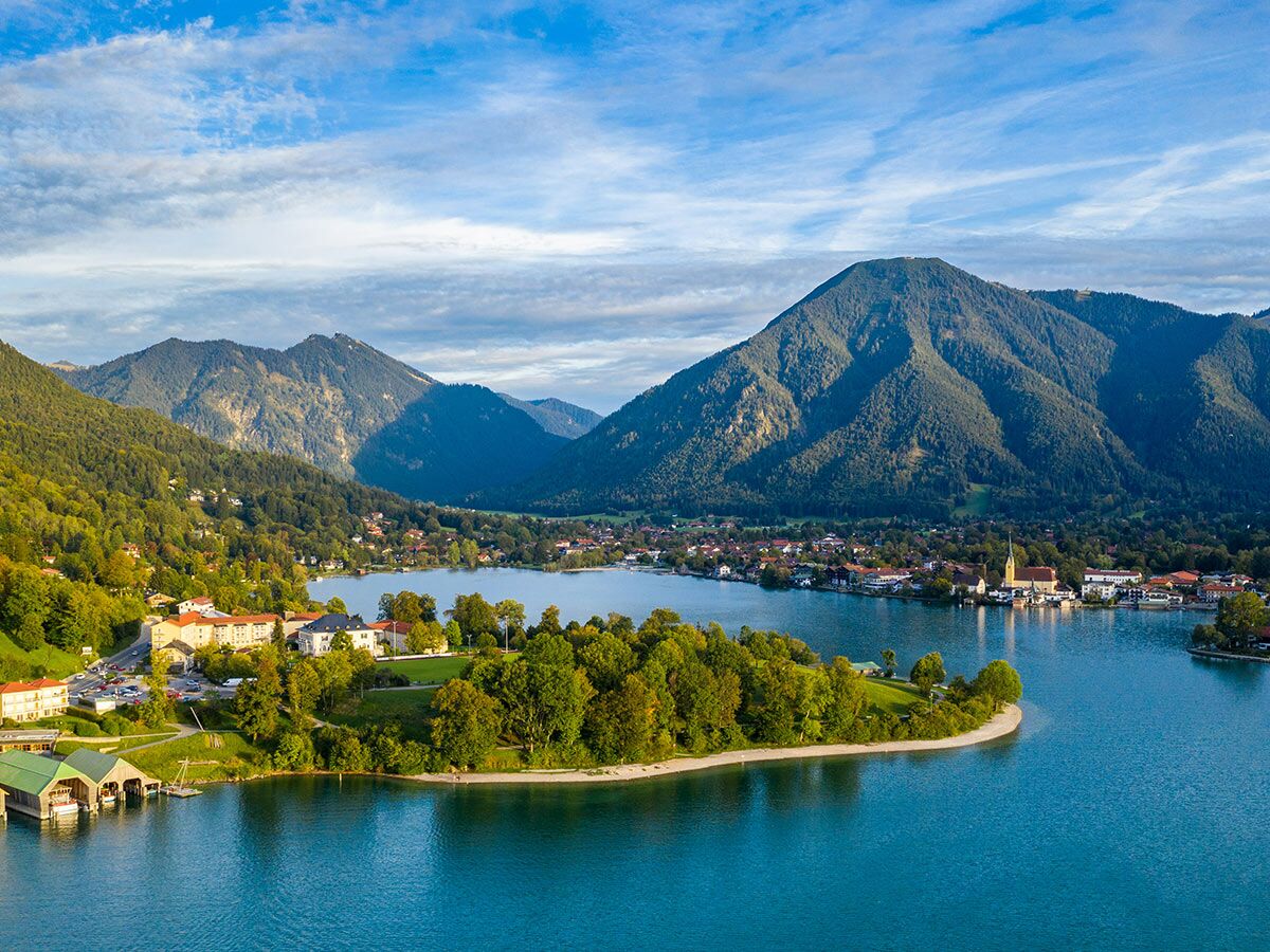 Malerischer Tegernsee in Bayern mit klarem blauen Wasser, bewaldeten Bergen, Bootshäusern und einem idyllischen Dorf.