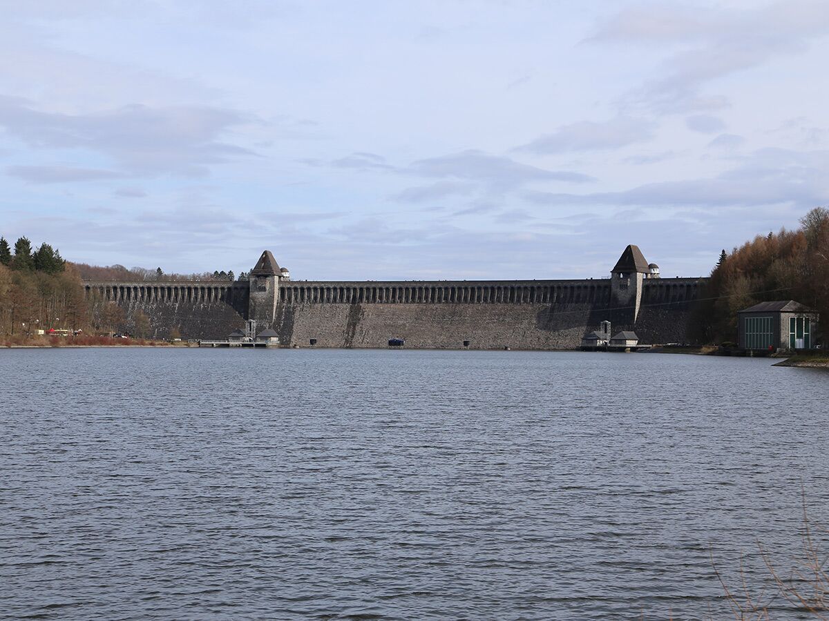 Blick über einen See auf eine imposante historische Staumauer aus Stein mit zwei markanten Türmen und bewaldeten Ufern unter bewölktem Himmel.