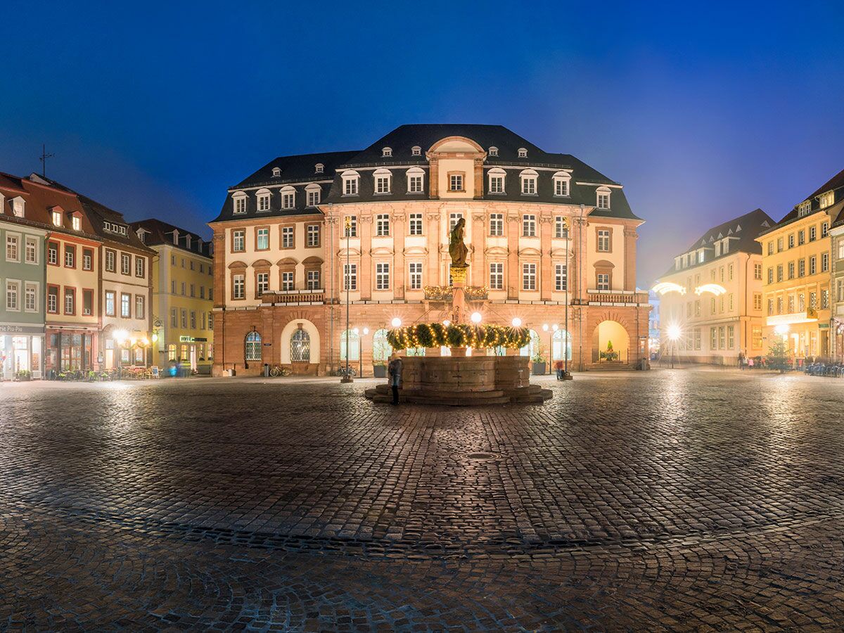 Historischer Marktplatz Heidelberg bei Abenddämmerung mit beleuchtetem Rathaus, Herkulesbrunnen und weihnachtlichen Lichtern auf Kopfsteinpflaster.