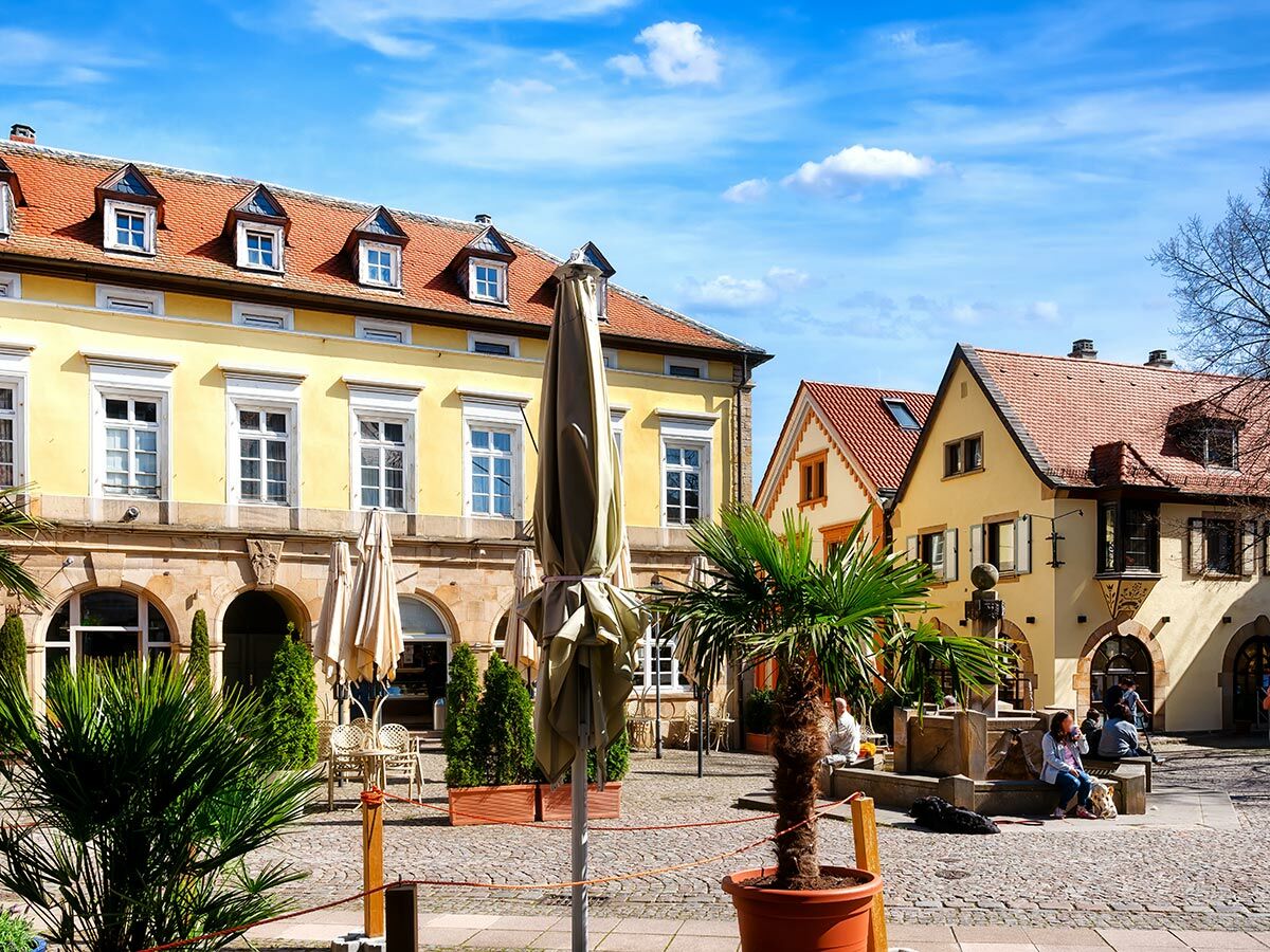 Historischer Marktplatz mit gelben Gebäuden, roten Dächern, Cafés, Menschen, Palmen und Brunnen unter blauem Himmel.