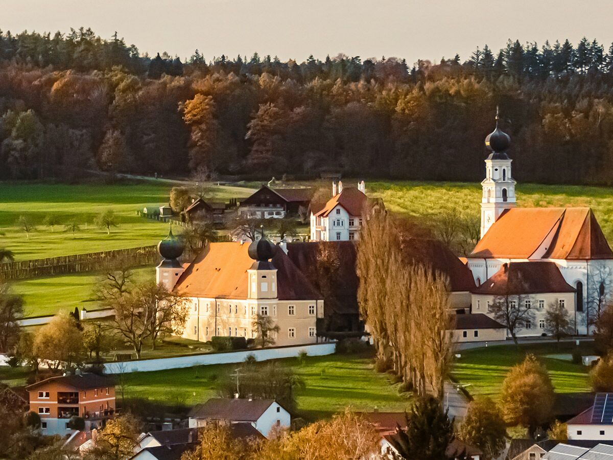 Barockes Kloster mit Kirche und Zwiebeltürmen in ländlicher Herbstlandschaft mit grünen Wiesen und Wäldern unter strahlender Sonne.