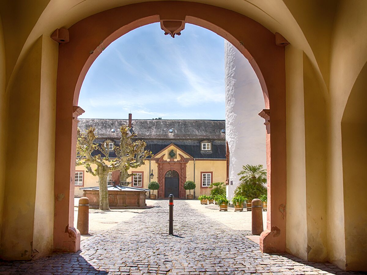 Blick durch einen roten Gewölbegang auf einen sonnigen historischen Innenhof mit Brunnen, altem Baum und gelbem Gebäude.