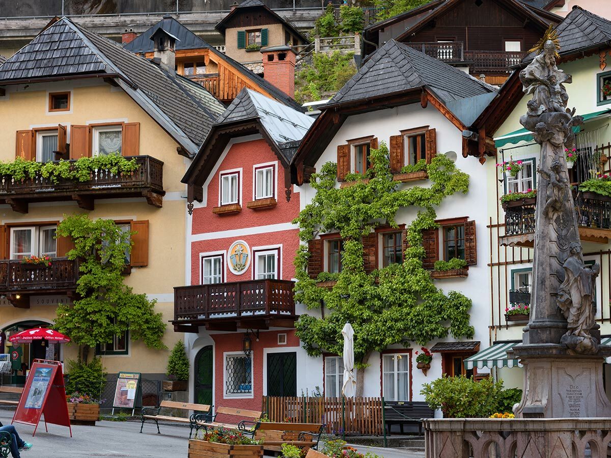 Blick auf farbenfrohe, traditionelle Häuser in Hallstatt, Österreich, mit Holzbalkonen, grünen Pflanzen und einer historischen Pestsäule.