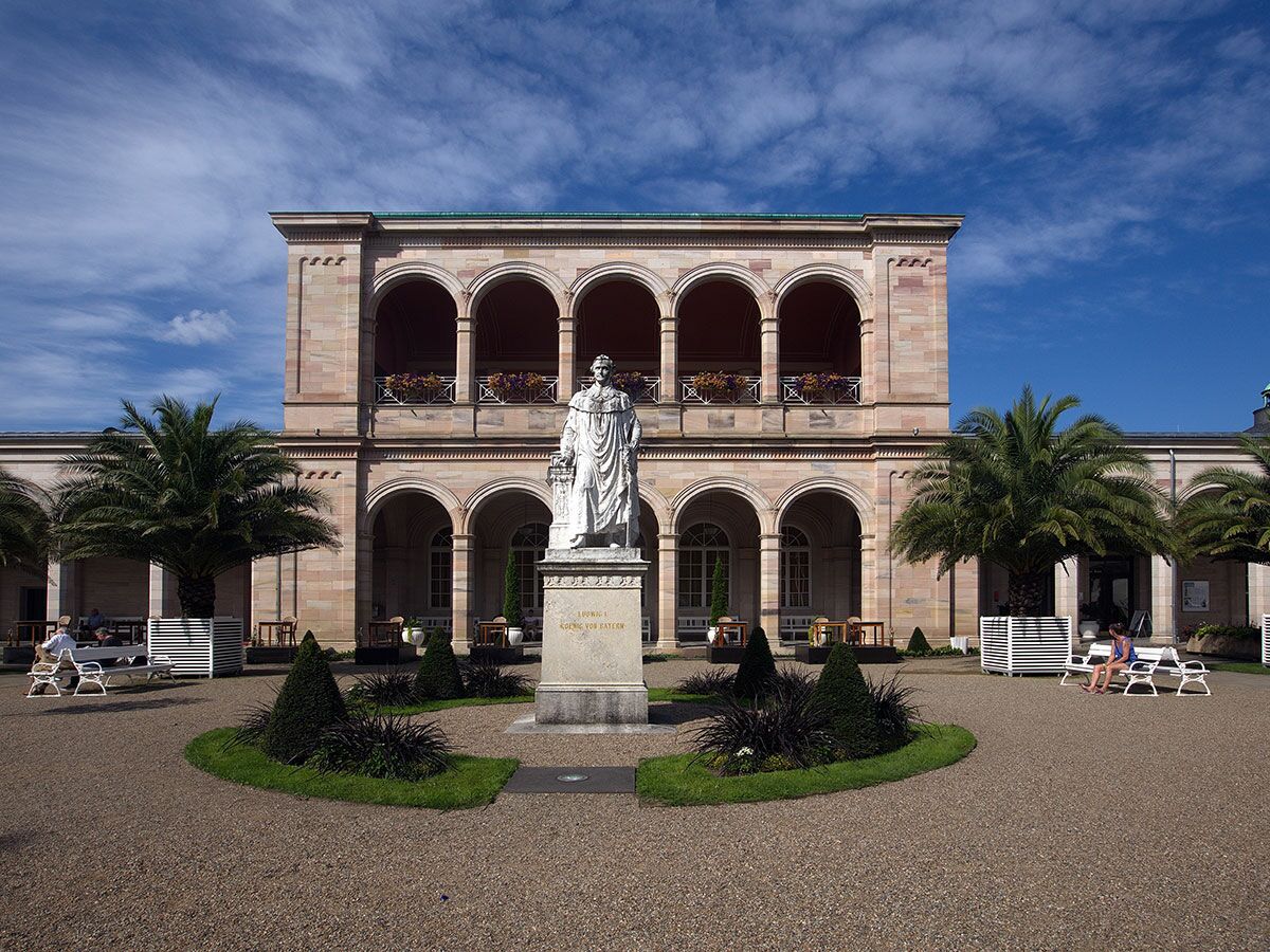 Außenansicht des Kurhauses Bad Kissingen mit Ludwig I Statue, Palmen, Bänken und Garten unter blauem Himmel.