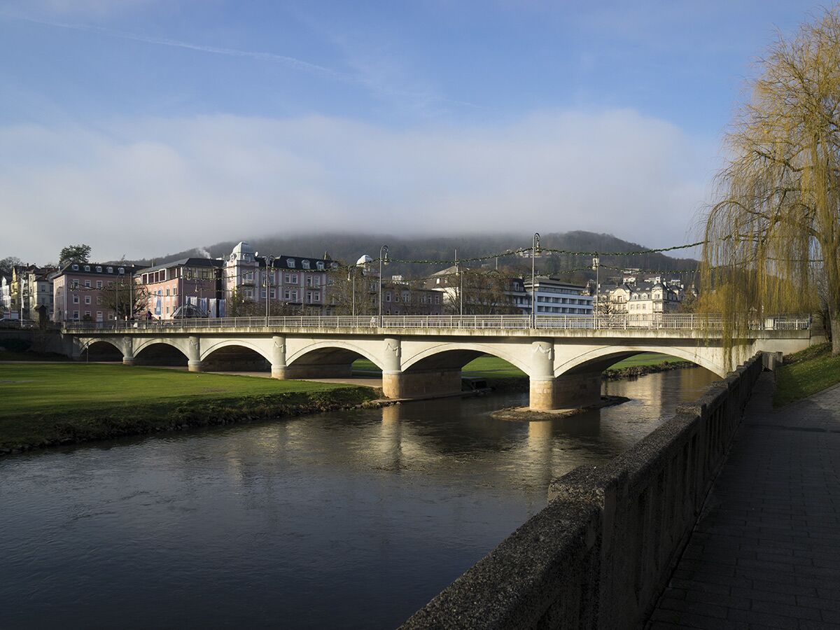 Panorama einer Stadt am Fluss mit mehrbögiger Brücke, grünen Ufern, bunten Gebäuden und nebelverhangenen Hügeln im Morgenlicht.