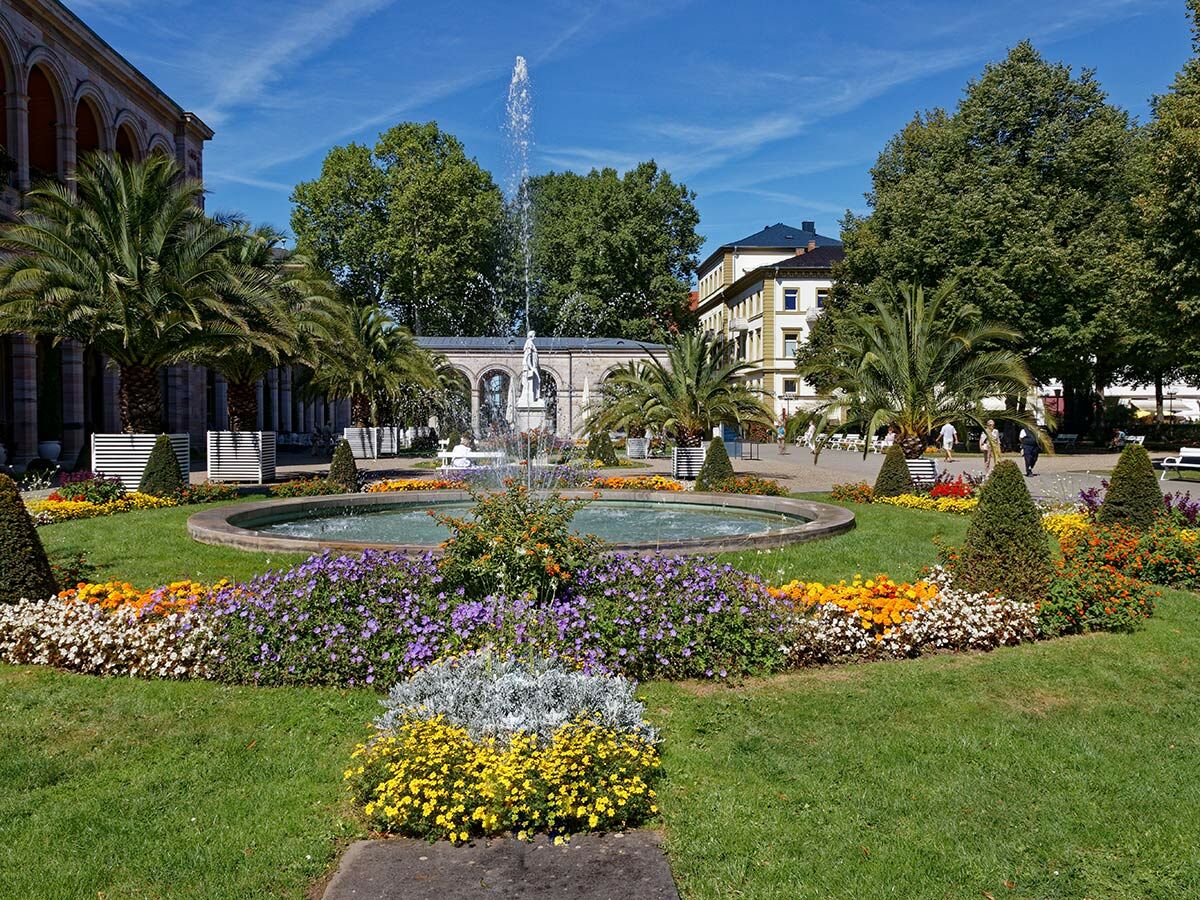 Sonnige Aufnahme eines Kurparks mit großem Springbrunnen, bunten Blumenbeeten, Palmen und historischen Gebäuden.