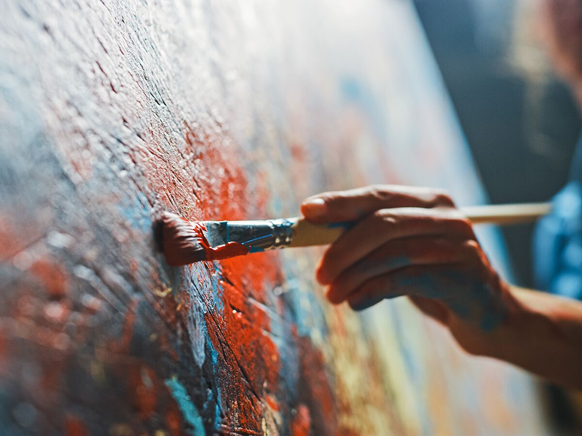 Nahaufnahme einer Hand, die mit Pinsel dicke rote und blaue Farbe auf strukturierte Leinwand aufträgt.