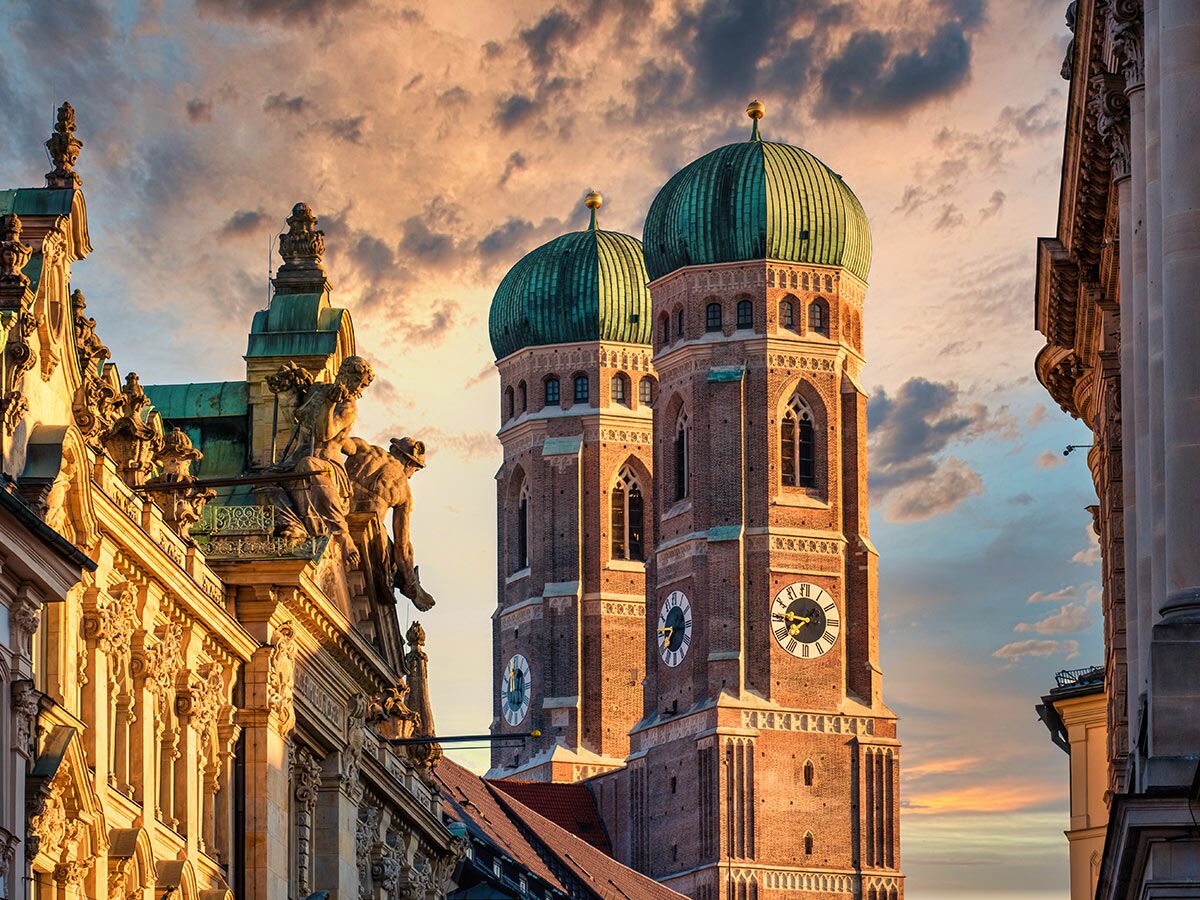 Frauenkirche München mit markanten Zwiebeltürmen im goldenen Licht des Sonnenuntergangs, umgeben von historischen Gebäuden.