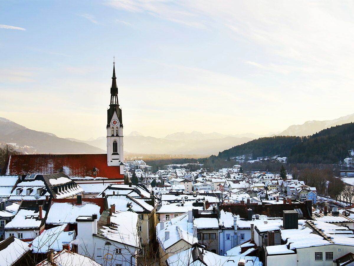 Panoramablick auf verschneite Alpenstadt mit markantem Kirchturm, roten Dächern und Bergen im Hintergrund bei Sonnenaufgang.