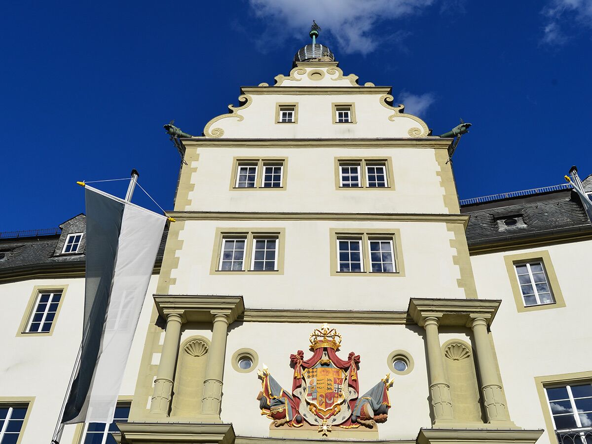 Historisches Gebäude mit verziertem Giebel, Wappen, Wasserspeiern und schwarz-weißer Fahne unter blauem Himmel.