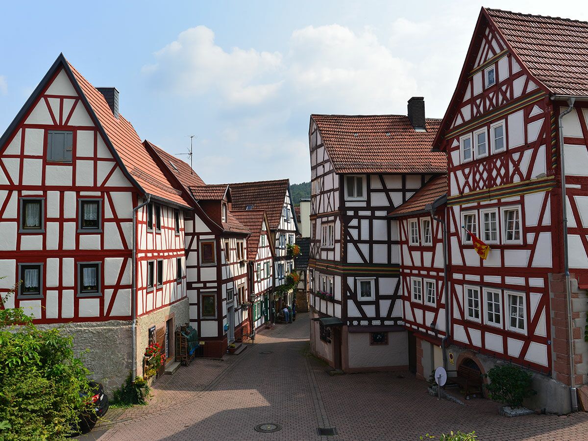 Reihe historischer Fachwerkhäuser mit roten Holzrahmen und roten Ziegeldächern entlang einer gepflasterten Dorfstraße.
