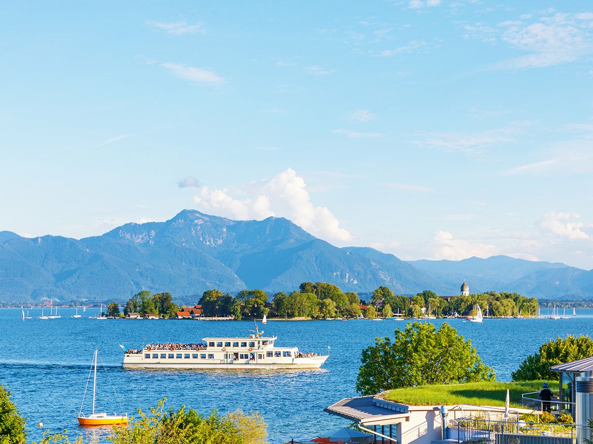 Panoramablick auf den Starnberger See mit Passagierschiff, bewaldeten Inseln, Segelbooten und Alpen im Hintergrund.