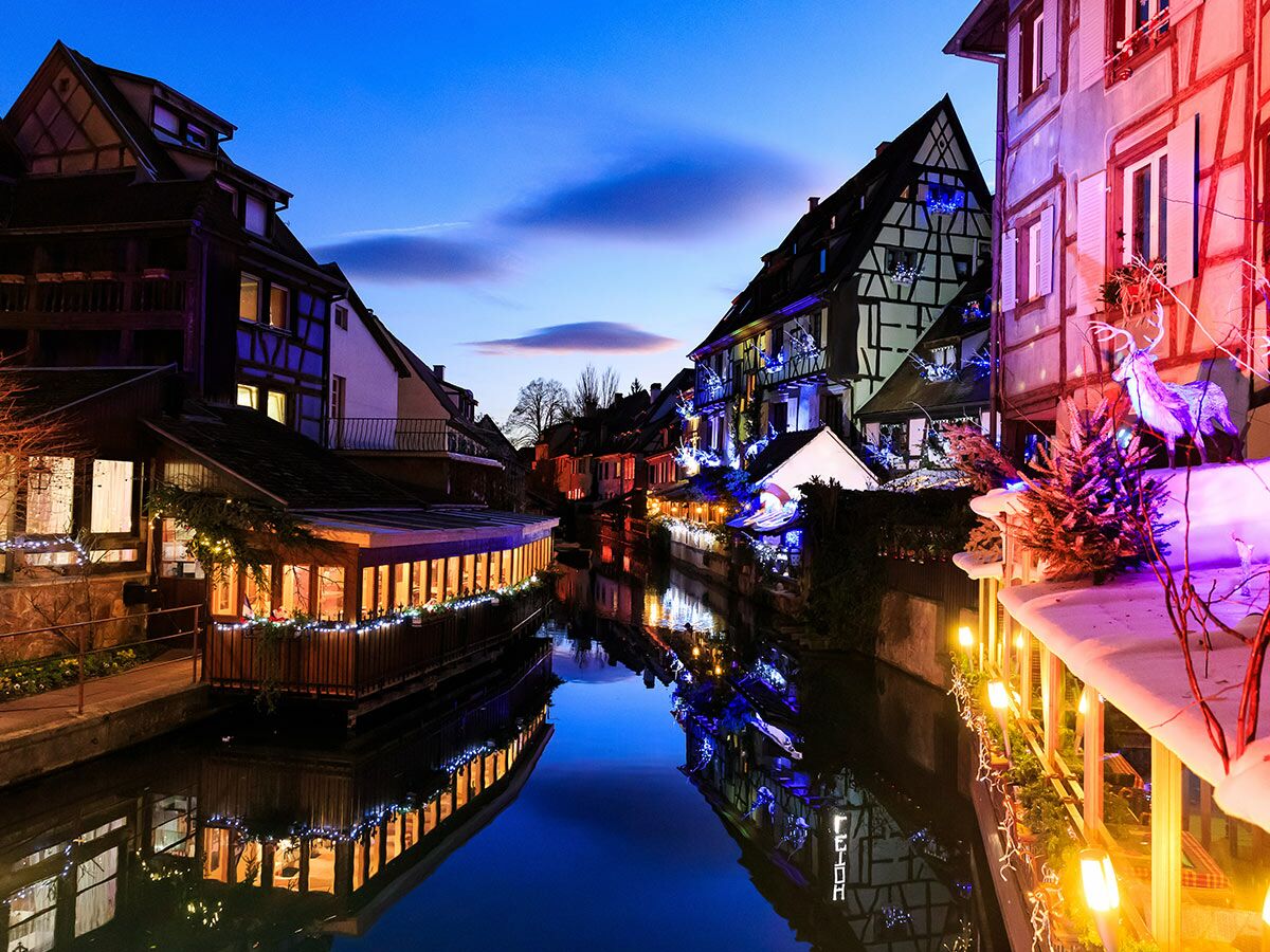 Kanal in Colmar mit festlich beleuchteten Fachwerkhäusern und Weihnachtsdekorationen bei Dämmerung, die sich im Wasser spiegeln.