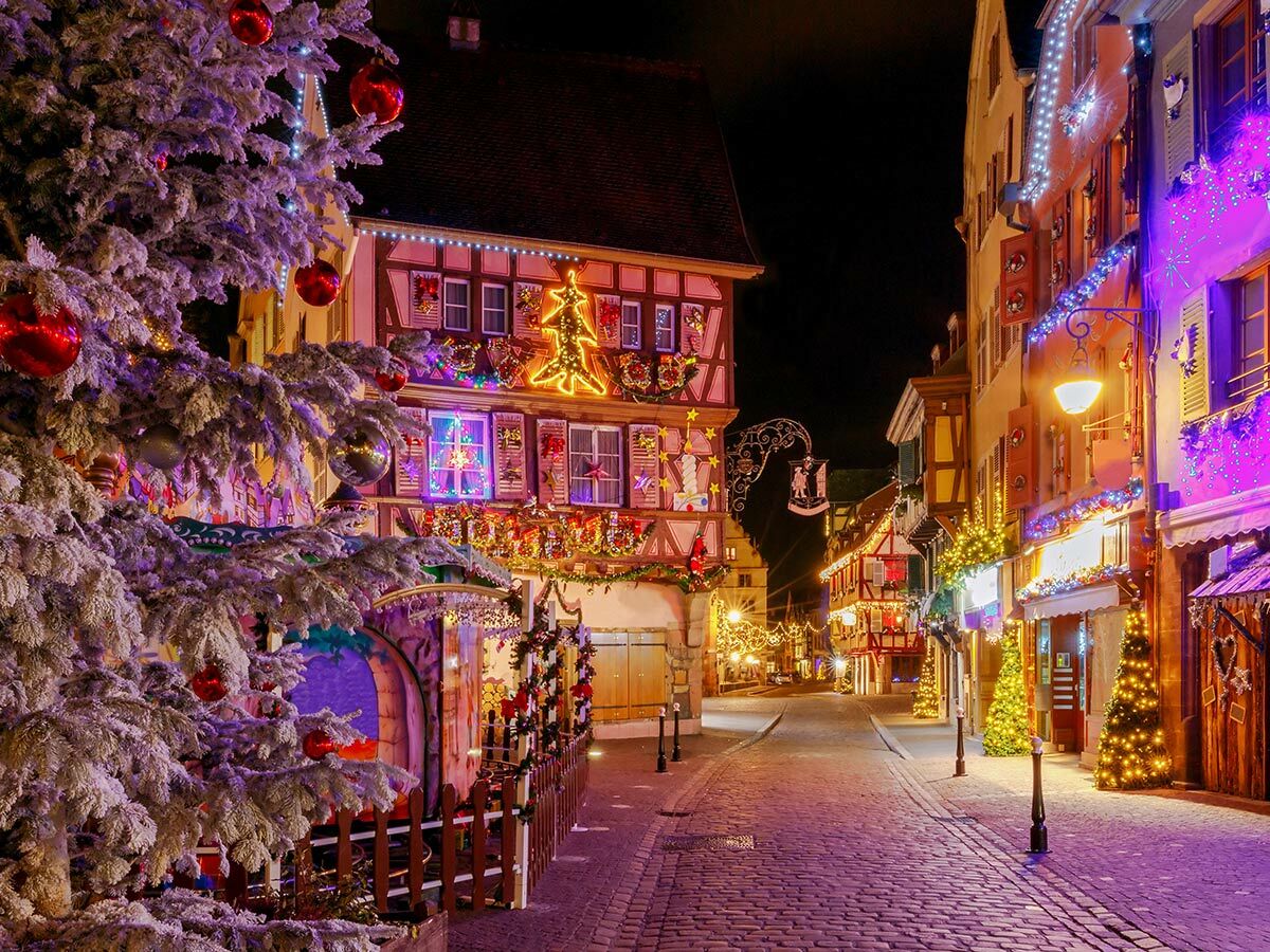 Festlich beleuchteter Weihnachtsmarkt bei Nacht in einer europäischen Altstadt mit Fachwerkhäusern, Lichterketten und einem großen Weihnachtsbaum.