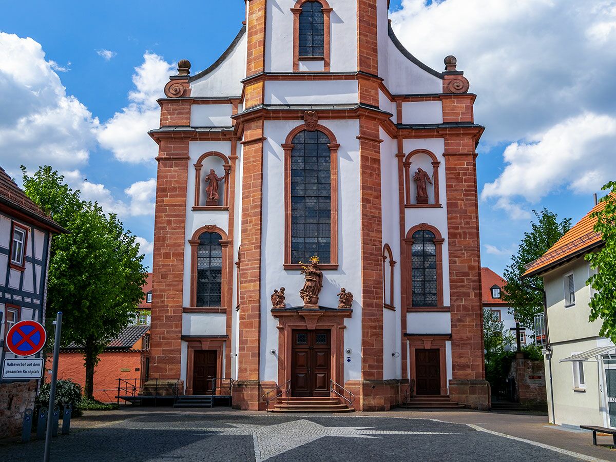 Historische Barockkirche mit roten Sandsteinelementen und weißen Wänden auf einem sonnigen Kirchplatz, umgeben von alten Häusern und Bäumen.