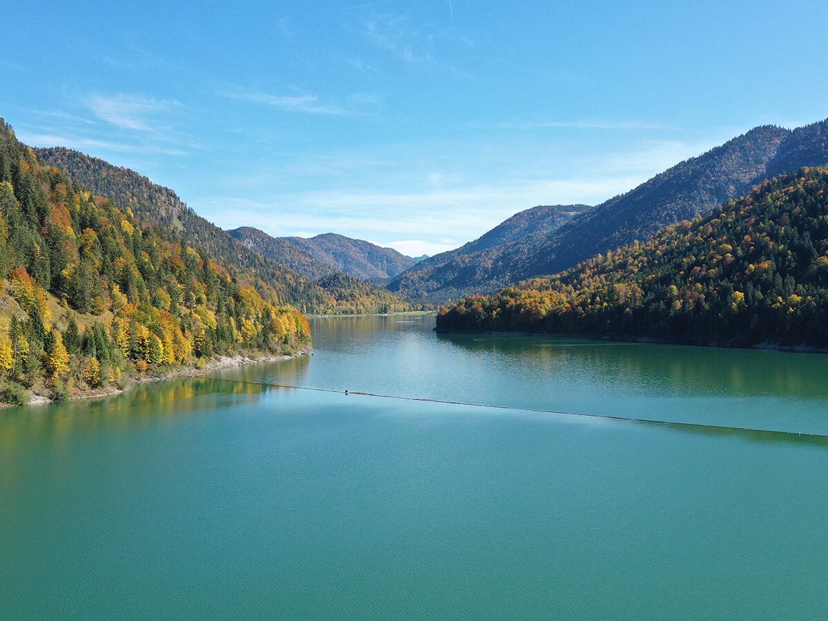 Panoramablick auf einen türkisblauen Bergsee umgeben von herbstlich gefärbten Wäldern und Bergen unter blauem Himmel.