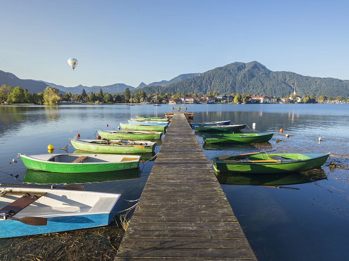 Idyllischer Tegernsee mit hölzernem Steg, grünen Ruderbooten und Heißluftballon über Berglanschaft und Dorf.