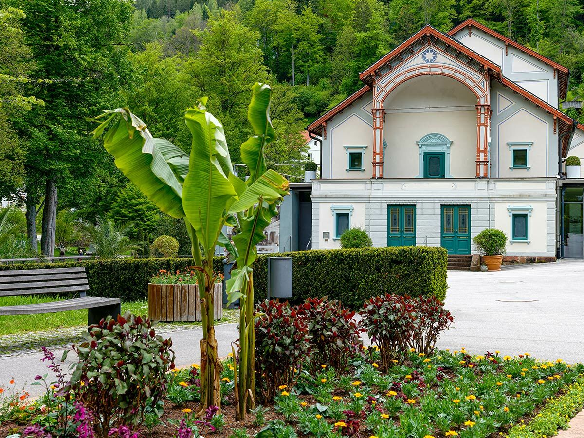 Elegantes, helles Gebäude mit Bogenportal und grünen Türen umgeben von einem üppigen Park mit Blumen und Bäumen.