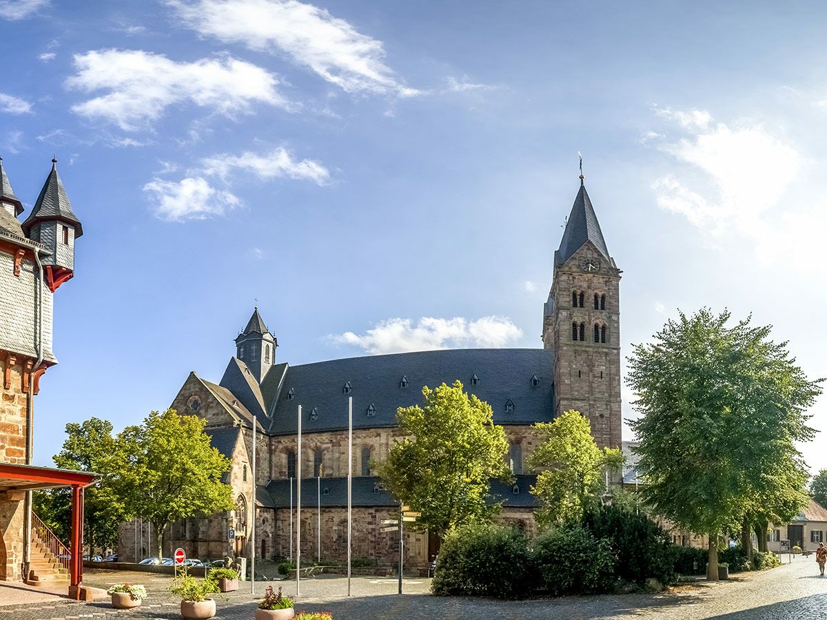 Blick auf eine historische, romanische Steinkirche mit hohem Glockenturm und Uhr an einem sonnigen Tag in einer deutschen Stadt.