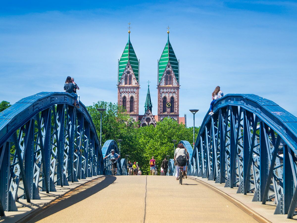 Menschen und Radfahrer auf der Blauen Brücke in Freiburg mit der St. Pauluskirche im Hintergrund an einem sonnigen Tag.