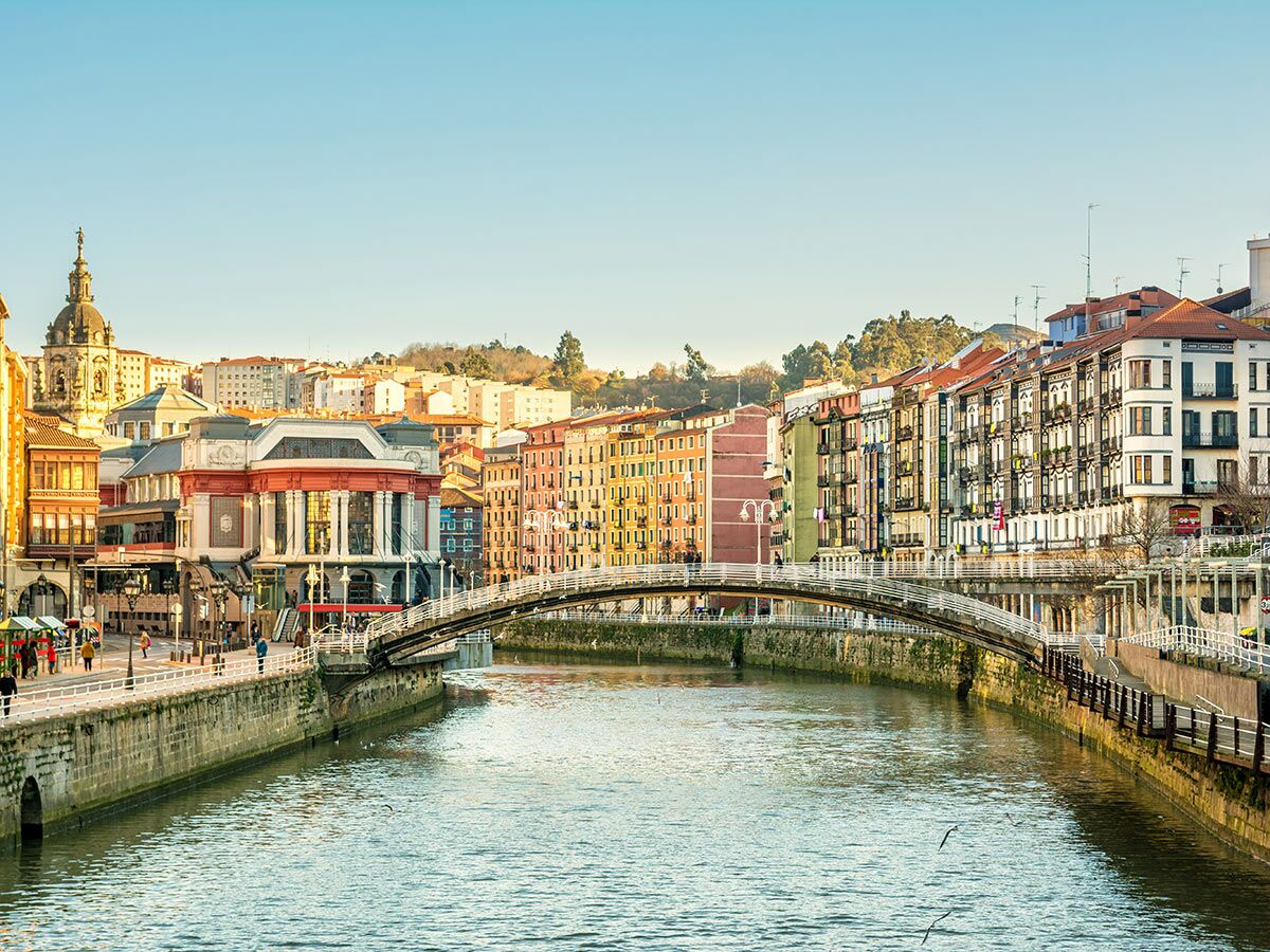 Panoramablick auf Bilbao mit dem Fluss Nervión, einer Bogenbrücke, historischen bunten Gebäuden und blauem Himmel.