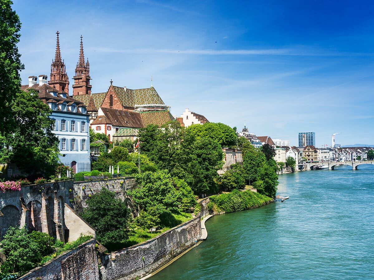 Historische Stadtansicht von Basel, Schweiz, mit dem imposanten roten Münster und dem blauen Rhein, umgeben von grünen Bäumen und Altstadtgebäuden.
