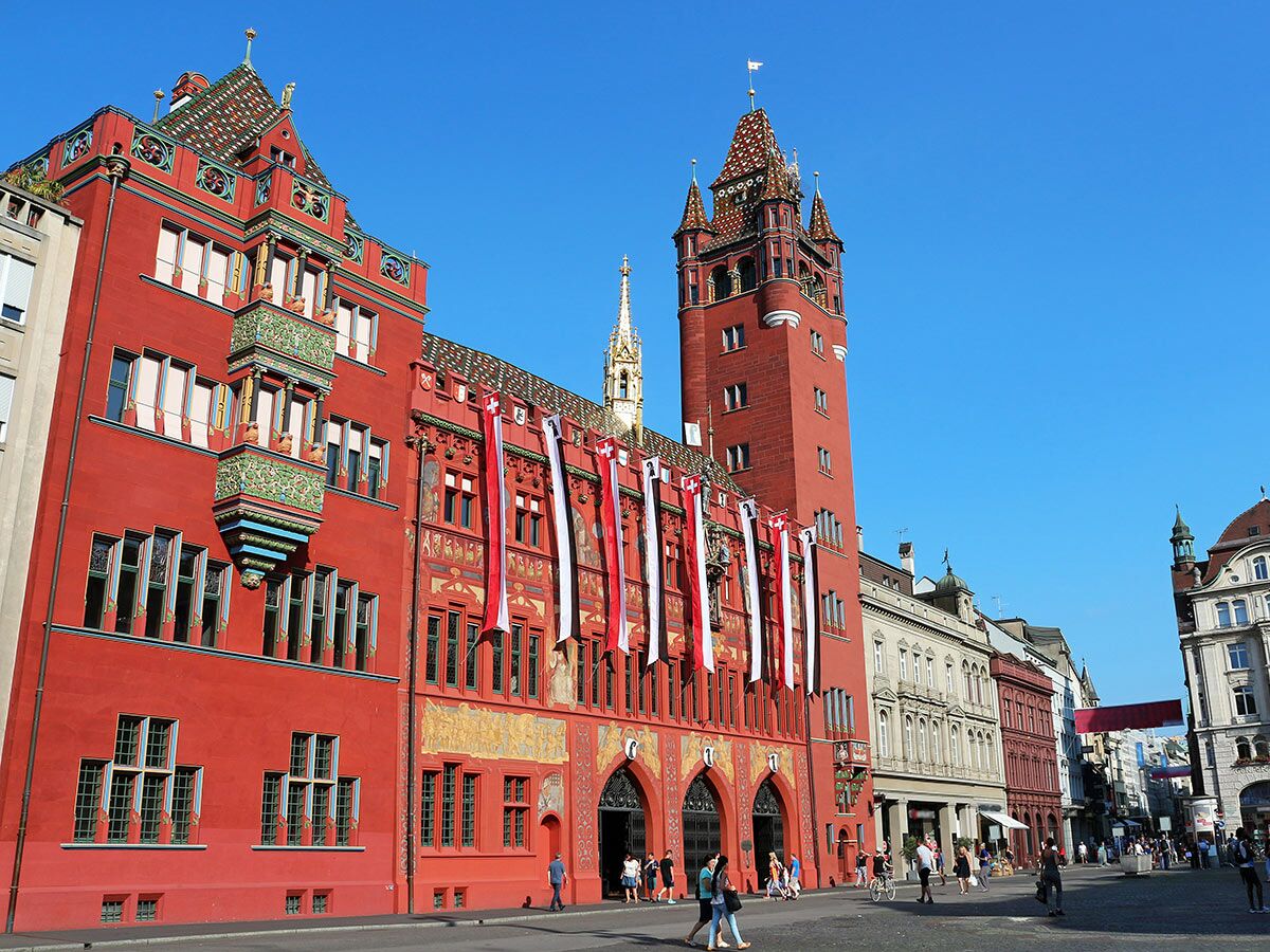 Rotes Rathaus Basel am Marktplatz unter blauem Himmel, mit Passanten und Schweizer Fahnen geschmückt.