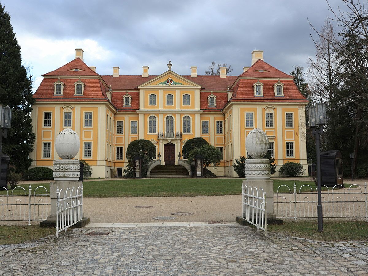 Großes, historisches Barockschloss mit gelber Fassade, rotem Dach und eleganten Eingangstoren an einem bewölkten Tag.