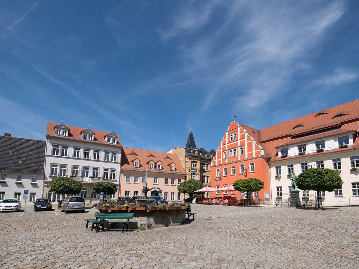Blick auf einen sonnigen, historischen Stadtplatz mit farbigen Gebäuden, Kopfsteinpflaster, Brunnen und Bänken in einer europäischen Altstadt.