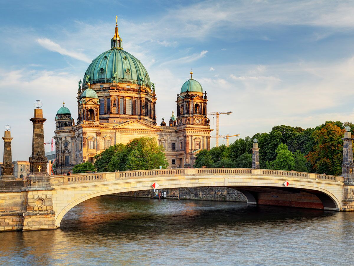 Majestätischer Berliner Dom bei Sonnenuntergang, mit Spree Fluss, Brücke, Bäumen und blauem Himmel.
