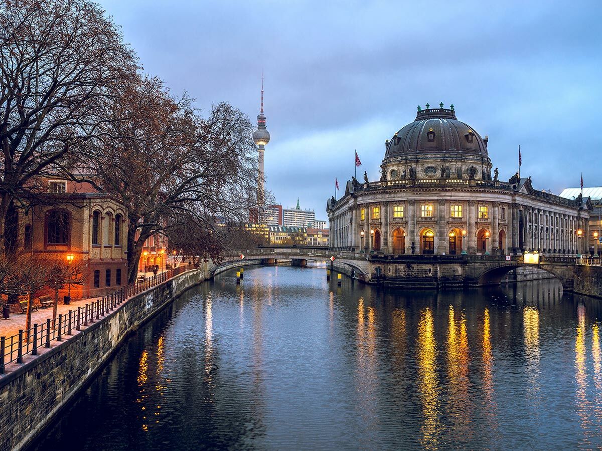 Abendliche Ansicht der Berliner Museumsinsel mit Bode-Museum und Fernsehturm an der Spree. Lichter spiegeln sich im Wasser.
