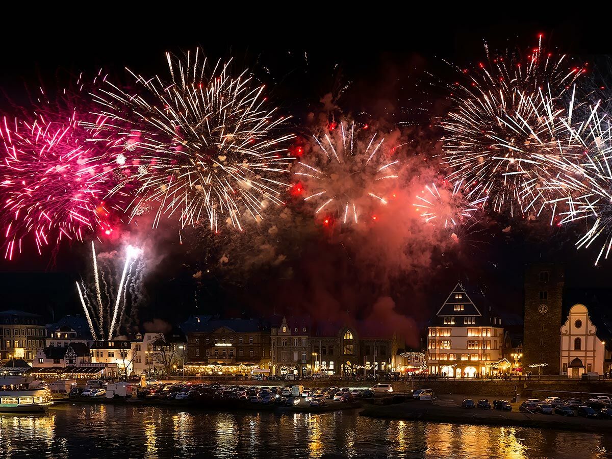 Spektakuläres Feuerwerk in Pink, Gold und Rot leuchtet über einer beleuchteten Flussstadt mit historischen Gebäuden.