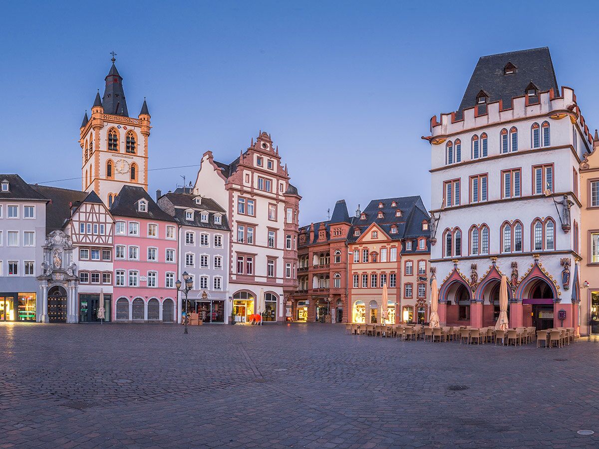 Blick auf den beleuchteten Hauptmarkt in Trier bei Abenddämmerung mit historischen Gebäuden, der St. Gangolf Kirche und leeren Caféterrassen.
