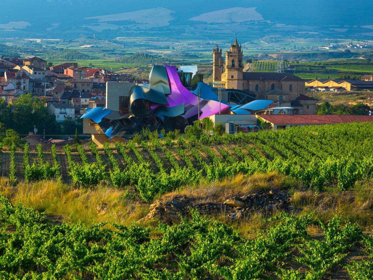 Modernes Hotel Marqués de Riscal in Elciego, Rioja, Spanien, umgeben von grünen Weinbergen, traditionellem Dorf und Kirche.