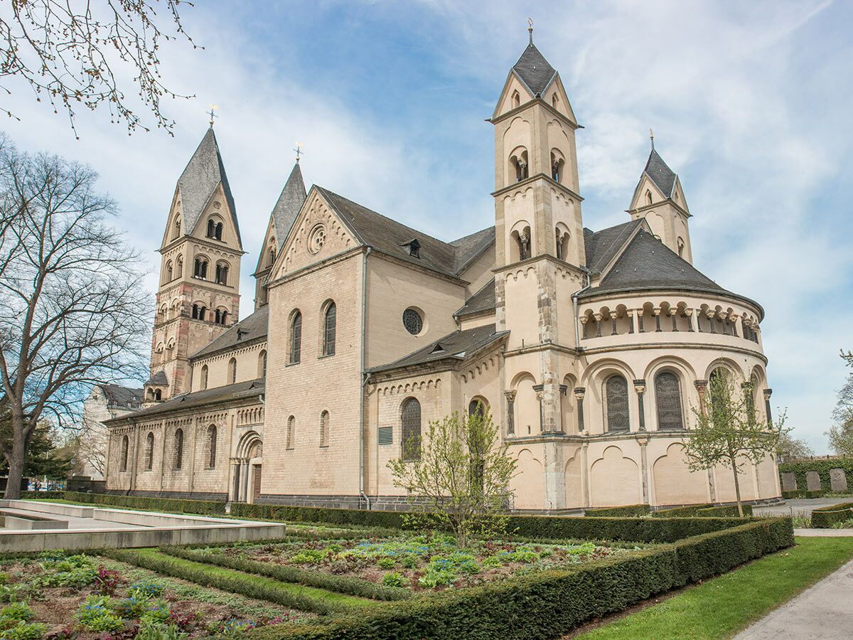 Ansicht der imposanten romanischen Basilika mit mehreren Türmen, umgeben von gepflegten Gärten an einem sonnigen Frühlingstag.