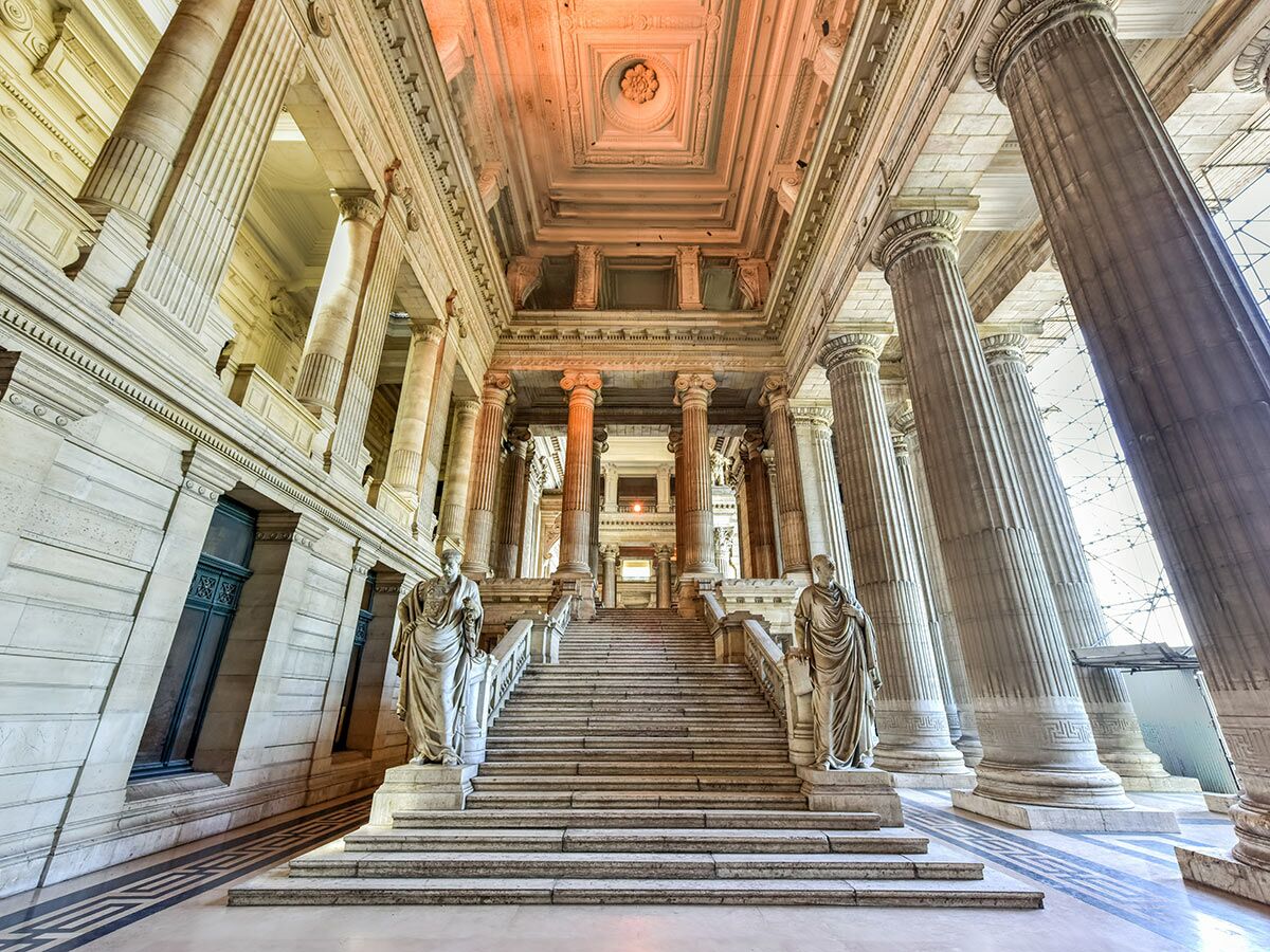 Innenansicht des Justizpalastes Brüssel mit monumentaler Treppe, hohen Säulen, Statuen und Deckenbeleuchtung in Steinfarben.