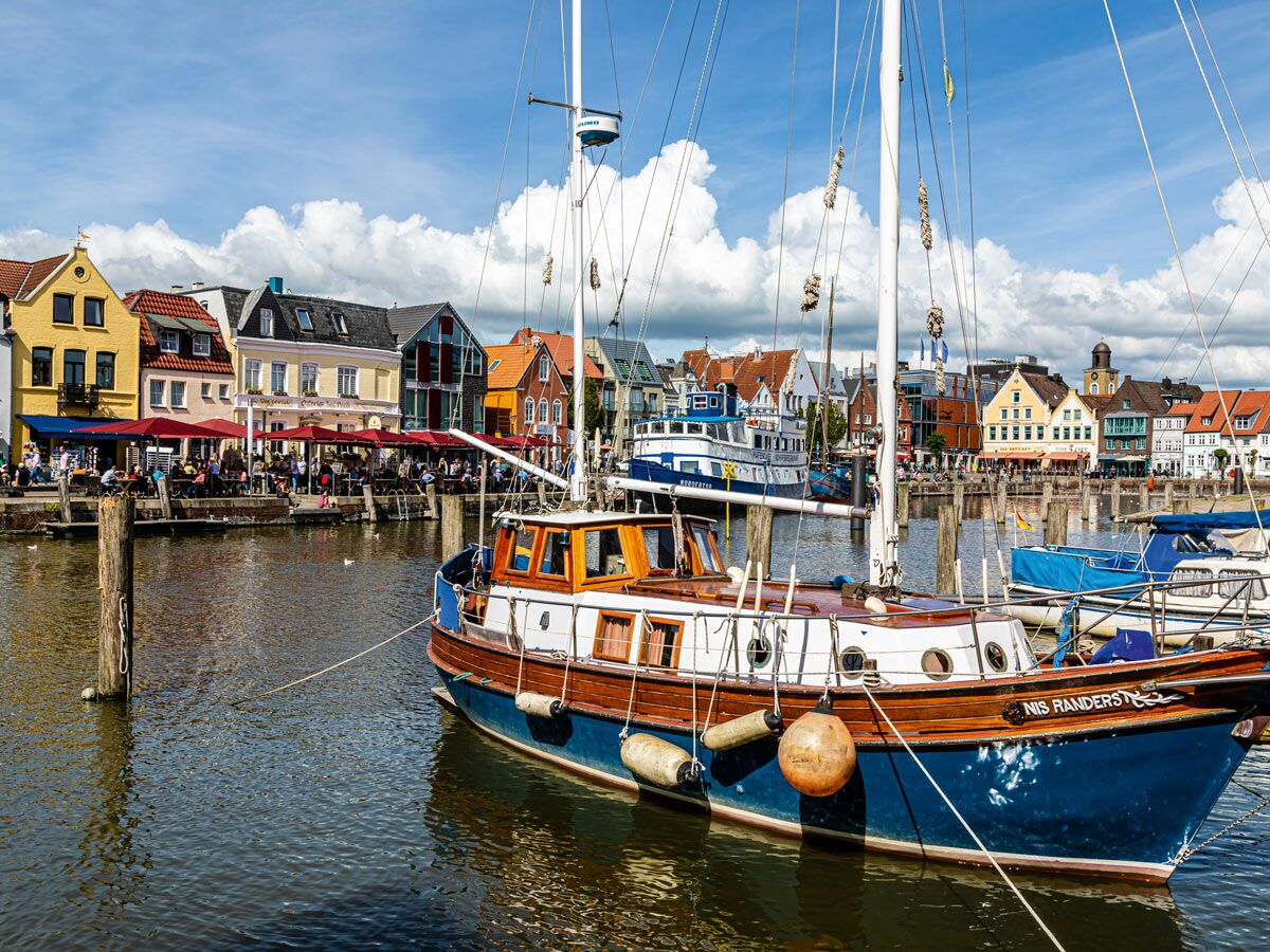 Blaues Segelboot Nis Randers ankert im Hafen von Büsum, belebte Promenade mit bunten Häusern unter blauem Himmel.
