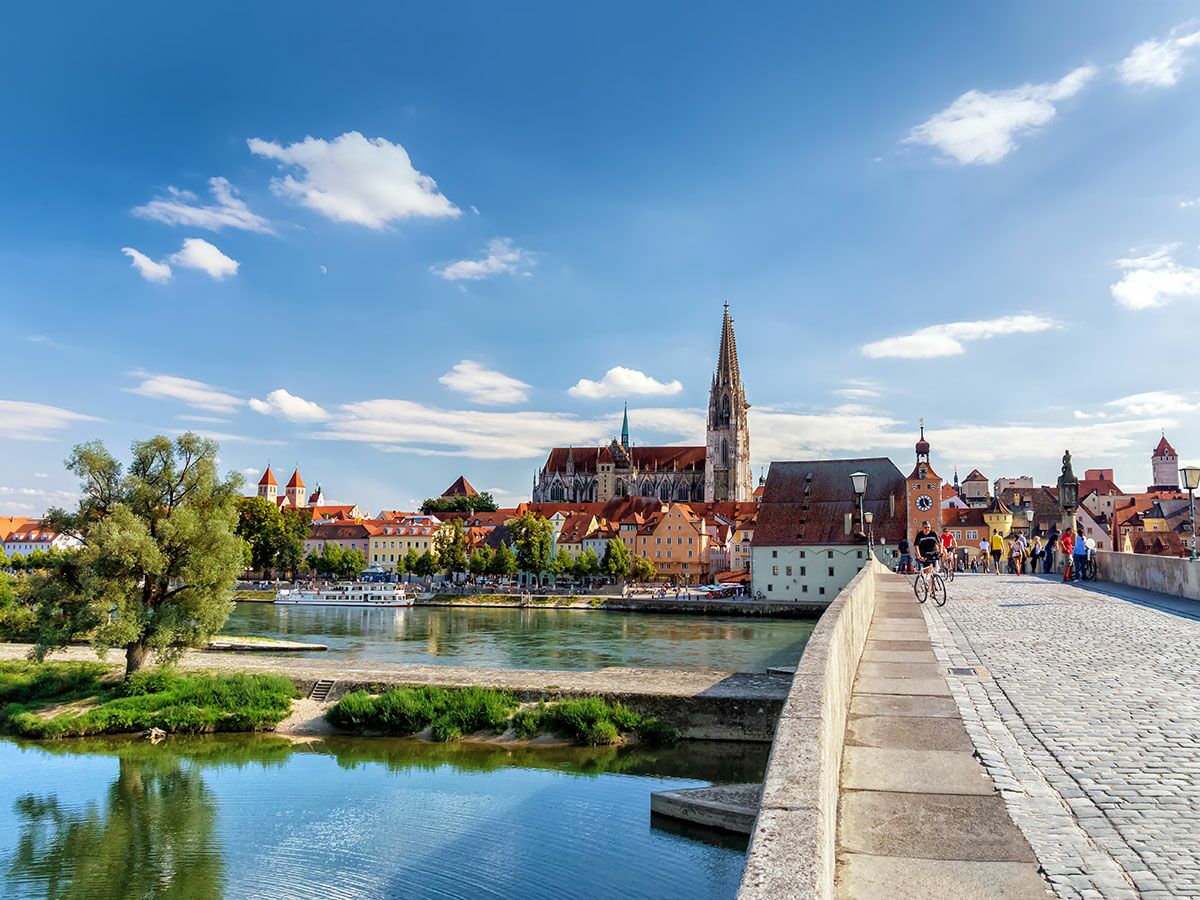 Blick auf Regensburg mit Steinerner Brücke, Donau, Dom St. Peter und historischer Altstadt unter blauem Himmel mit Wolken.