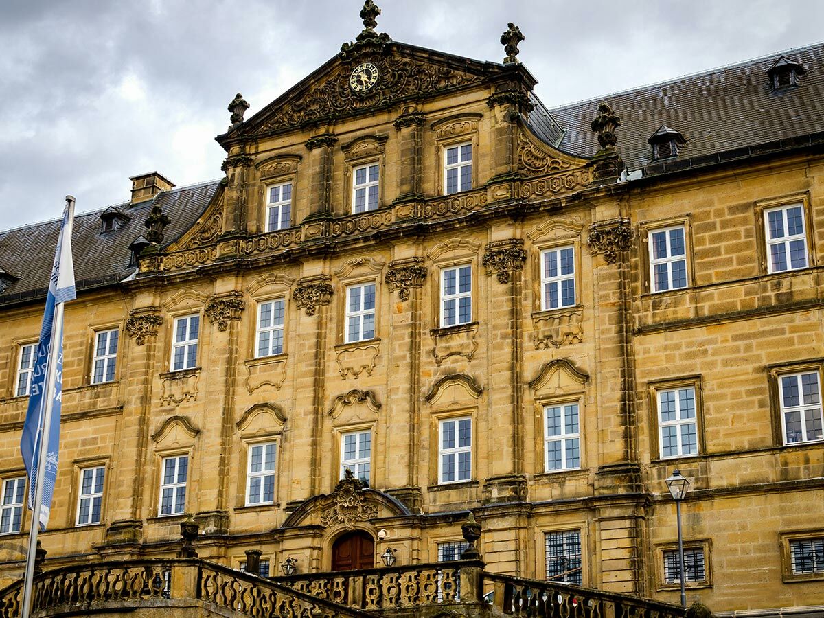 Historische Neue Residenz Bamberg mit barocker Fassade, vielen Fenstern und blauer Flagge unter bewölktem Himmel.