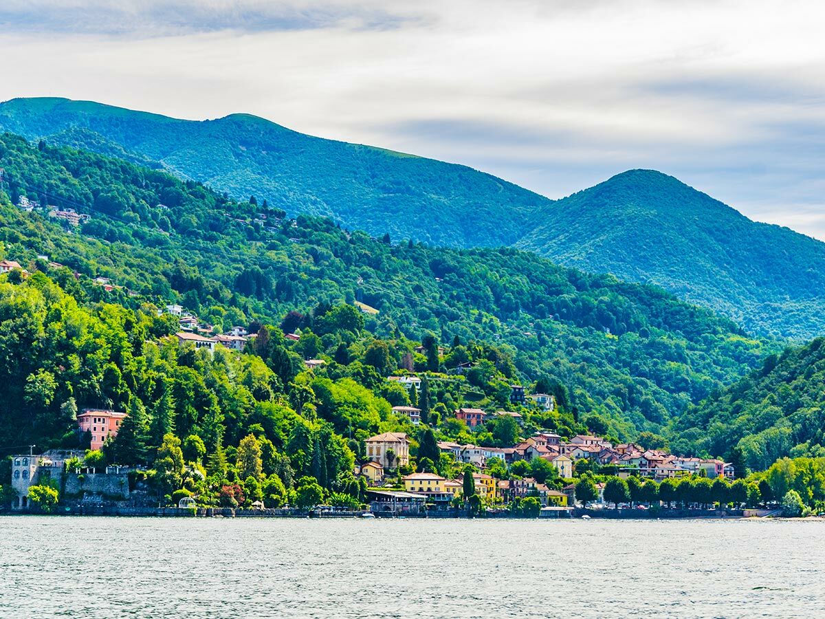 Blick über einen ruhigen See auf grüne, bewaldete Berge, an deren Hängen sich charmante Dörfer mit bunten Häusern und Villen schmiegen. Italienische Landschaft.
