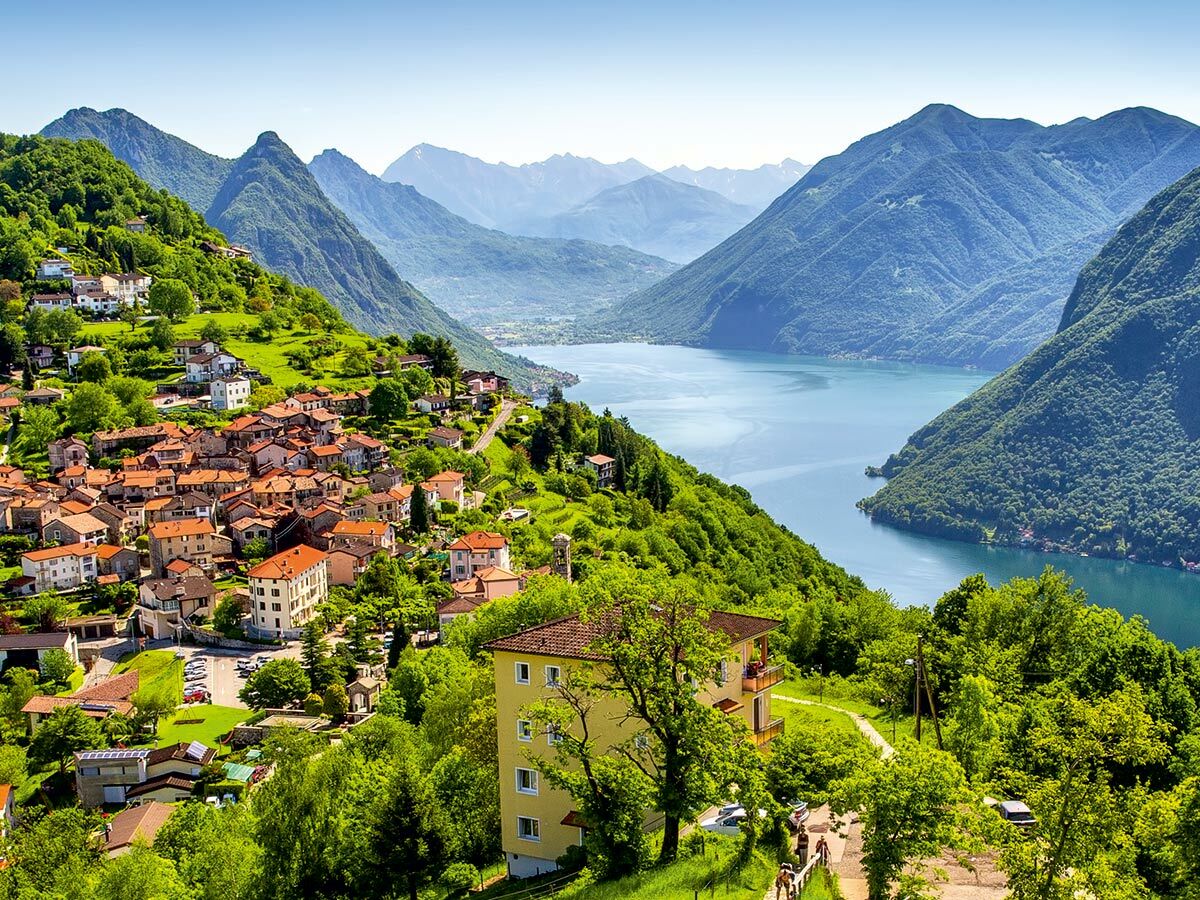 Panoramablick auf ein malerisches Bergdorf mit roten Dächern am Ufer eines blauen Sees, umgeben von grünen Bergen unter klarem Himmel.
