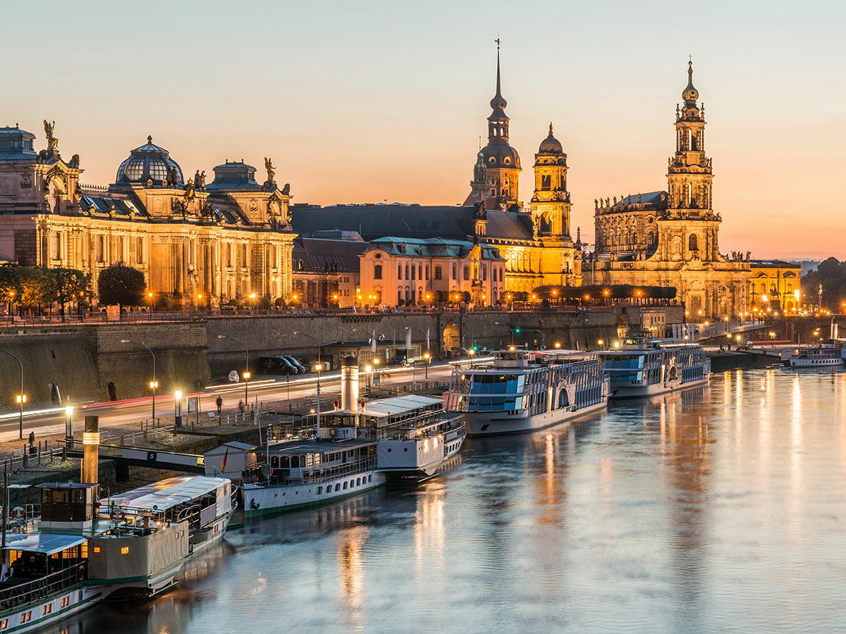 Panoramablick auf das historische Dresden bei Dämmerung, beleuchtete Gebäude am Elbufer und Schiffe auf dem Fluss.